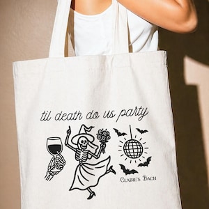 Op de afbeelding: Een witte canvas draagtas met een zwarte grafische print van een skelet in een jurk en hoed die een boeket, een wijnglas, een discobal en vleermuizen vasthoudt. De tekst luidt "til death do us party" en "Claire's BACH" in zwarte letters.