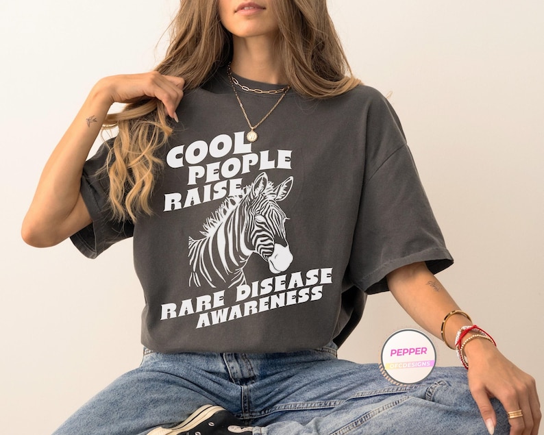 K&ouml;nnte beinhalten: Ein anthrazitfarbenes T-Shirt mit einem wei&szlig;en Zebra-Motiv und dem Text "Cool People Raise Rare Disease Awareness".