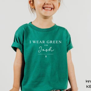 May include: A child wearing a teal green t-shirt with the text "I WEAR GREEN FOR Josh" in white. Below the name is the text "CEREBRAL PALSY AWARENESS" and a ribbon graphic. The shirt has rolled-up sleeves.