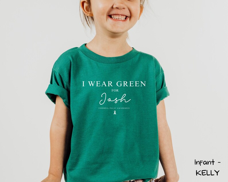 May include: A child wearing a teal green t-shirt with the text "I WEAR GREEN FOR Josh" in white. Below the name is the text "CEREBRAL PALSY AWARENESS" and a ribbon graphic. The shirt has rolled-up sleeves.