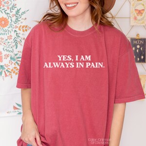 K&ouml;nnte beinhalten: Ein T-Shirt in einem kr&auml;ftigen Rotton mit dem in Wei&szlig; gedruckten Text "YES, I AM ALWAYS IN PAIN."