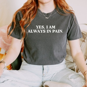 K&ouml;nnte beinhalten: Eine Frau tr&auml;gt ein dunkelgraues T-Shirt mit dem Aufdruck "YES, I AM ALWAYS IN PAIN." in wei&szlig;. Sie tr&auml;gt eine blaue Jeans und hat lange rote Haare.