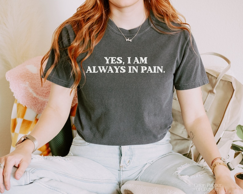 K&ouml;nnte beinhalten: Eine Frau tr&auml;gt ein dunkelgraues T-Shirt mit dem Aufdruck "YES, I AM ALWAYS IN PAIN." in wei&szlig;. Sie tr&auml;gt eine blaue Jeans und hat lange rote Haare.