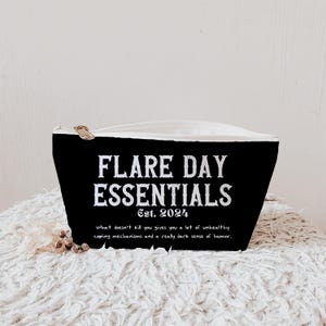 Pode incluir: Bolsa preta com z&iacute;per com o texto "FLARE DAY ESSENTIALS Est. 2024". A bolsa tamb&eacute;m tem a cita&ccedil;&atilde;o "O que n&atilde;o te mata te d&aacute; muitos mecanismos de enfrentamento pouco saud&aacute;veis e um senso de humor realmente sombrio."