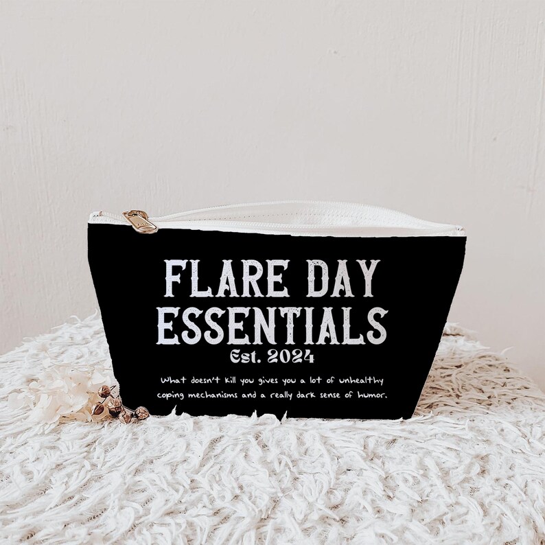 Pode incluir: Bolsa preta com z&iacute;per com o texto "FLARE DAY ESSENTIALS Est. 2024". A bolsa tamb&eacute;m tem a cita&ccedil;&atilde;o "O que n&atilde;o te mata te d&aacute; muitos mecanismos de enfrentamento pouco saud&aacute;veis e um senso de humor realmente sombrio."