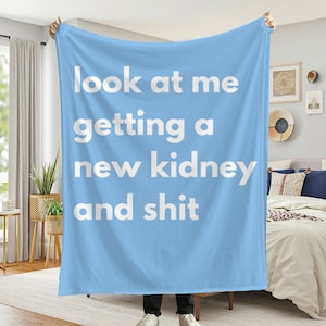 May include: A pale blue blanket featuring the white text: "look at me getting a new kidney and shit". The blanket is displayed in a room with a bed and a window.