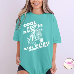 K&ouml;nnte beinhalten: T-Shirt in T&uuml;rkisblau mit einem wei&szlig;en Zebra-Motiv und dem Text "Cool People Raise Rare Disease Awareness."