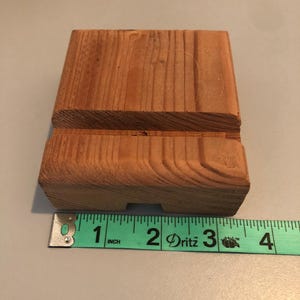 May include: A small wooden stand made of natural wood with a light brown color. The stand is approximately 2 inches wide and 2 inches long. It has a slot in the middle for holding a phone or other small object.