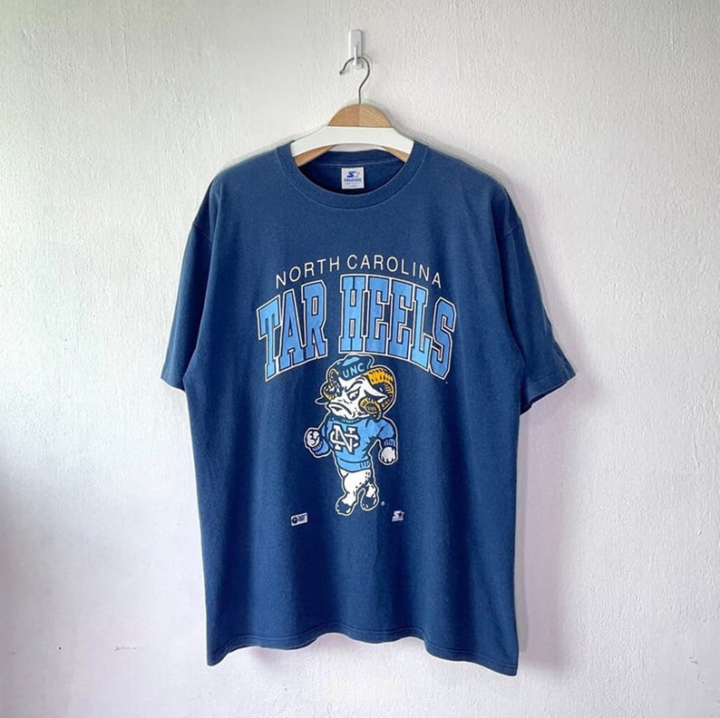 以下が含まれることがあります： ノースカロライナ・ターヒールズのロゴが入ったヴィンテージの青いTシャツ。ターヒールズのロゴは、青と白のユニフォームを着た雄羊のキャラクターで、その上に「NORTH CAROLINA TAR HEELS」の文字が書かれています。
