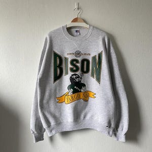 May include: A heather gray sweatshirt featuring the word "BISON" in green and white, with a graphic of a bison smoking a cigar. The sweatshirt also includes the text "NORTH DAKOTA STATE" and "FARGO ND".