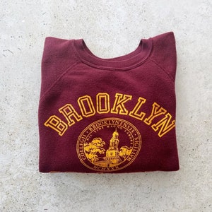 May include: A maroon sweatshirt with a gold embroidered design. The design features the word "Brooklyn" in a large arc above a circular seal with the words "Collegii Brooklynenis" and "Mcmx" in gold.