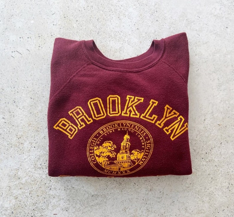 May include: A maroon sweatshirt with a gold embroidered design. The design features the word "Brooklyn" in a large arc above a circular seal with the words "Collegii Brooklynenis" and "Mcmx" in gold.