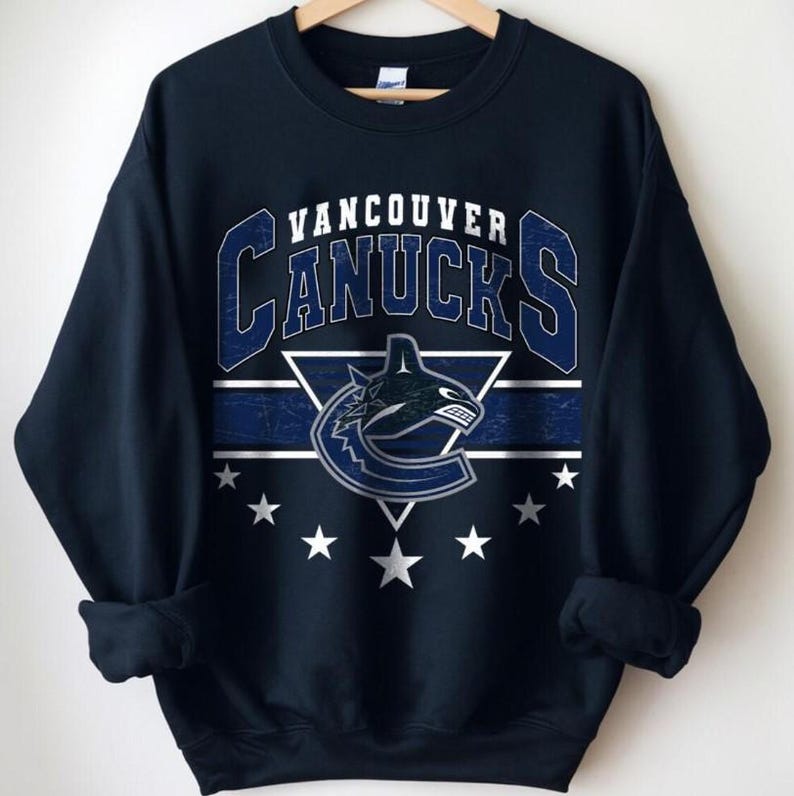 Pu&ograve; includere: Felpa blu navy con la scritta "Vancouver Canucks" in bianco e blu. Il design include il logo della squadra e stelle. La felpa &egrave; appesa a una gruccia di legno.