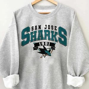 May include: A light gray sweatshirt with the text "SAN JOSE SHARKS 1991" in teal and black. The sweatshirt features the Sharks logo, a shark with hockey sticks. The sleeves are cuffed.