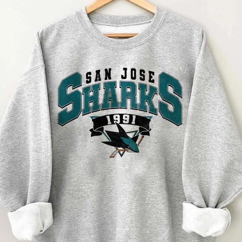 May include: A light gray sweatshirt with the text "SAN JOSE SHARKS 1991" in teal and black. The sweatshirt features the Sharks logo, a shark with hockey sticks. The sleeves are cuffed.