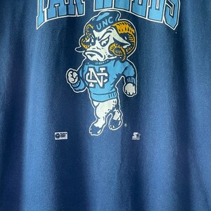 以下が含まれることがあります： 青いTシャツに、帽子に「UNC」、セーターに「N」と描かれた青いセーターを着た、漫画の羊のヴィンテージスタイルのグラフィックが描かれています。「RAMS」という文字が羊の上に、「STARTER」という文字が羊の下にあります。