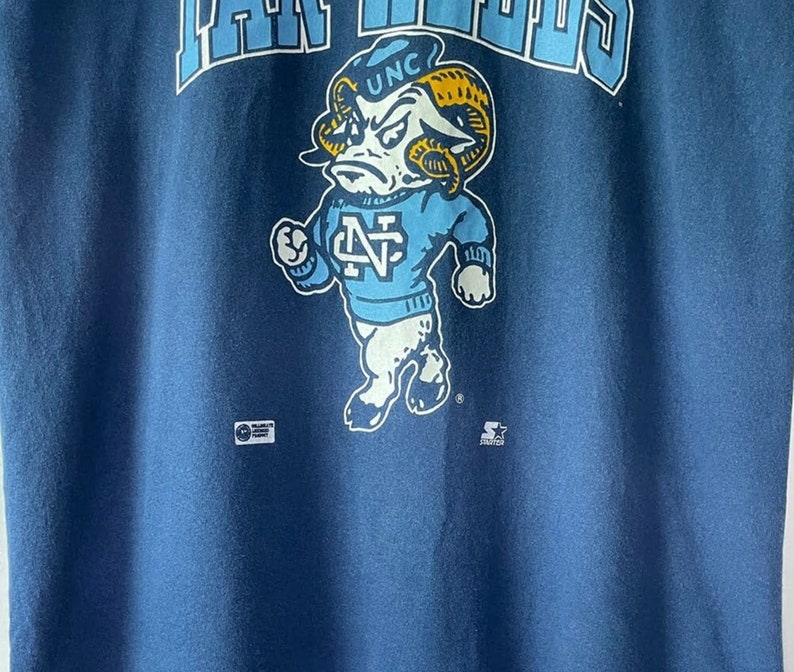 以下が含まれることがあります： 青いTシャツに、帽子に「UNC」、セーターに「N」と描かれた青いセーターを着た、漫画の羊のヴィンテージスタイルのグラフィックが描かれています。「RAMS」という文字が羊の上に、「STARTER」という文字が羊の下にあります。