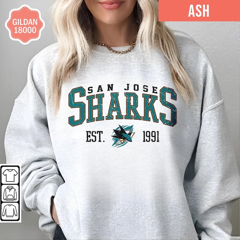 May include: A gray sweatshirt with the words "San Jose Sharks" in green and gold lettering. The text "Est. 1991" is below the team name. A teal and white shark logo is between the text.