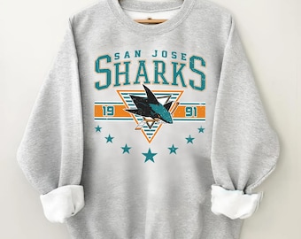 May include: A light gray sweatshirt with the text "SAN JOSE SHARKS" in teal and orange. The design includes a shark graphic, the numbers "19" and "91", and teal stars. The sweatshirt has rolled-up sleeves.