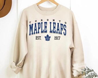 May include: A beige sweatshirt with the text "TORONTO MAPLE LEAFS EST. 1917" in navy blue. The sweatshirt features a small blue maple leaf logo.