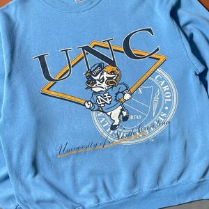 K&ouml;nnte beinhalten: Ein hellblaues Sweatshirt mit einem Vintage-Logo der UNC Tar Heels. Das Logo zeigt ein Widder-Maskottchen mit dem Text "UNC" dar&uuml;ber und "University of North Carolina" darunter.