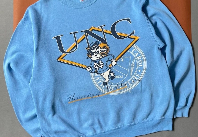 K&ouml;nnte beinhalten: Ein hellblaues Sweatshirt mit einem Vintage-Logo der UNC Tar Heels. Das Logo zeigt ein Widder-Maskottchen mit dem Text "UNC" dar&uuml;ber und "University of North Carolina" darunter.