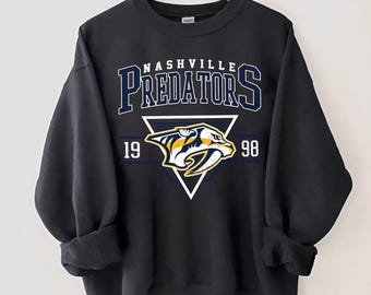 May include: Black sweatshirt with a white triangle and the words "Nashville Predators" in blue and yellow. The triangle contains a yellow and blue predator logo with the years "19" and "98" on either side.