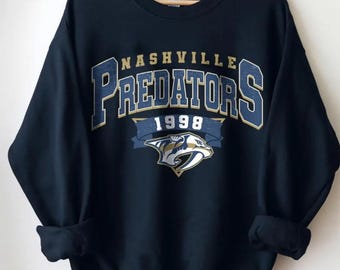 May include: Navy blue sweatshirt featuring the text "Nashville Predators" in a bold, arched design with gold and blue accents. The year "1998" is displayed below a banner, and the team logo is at the bottom.