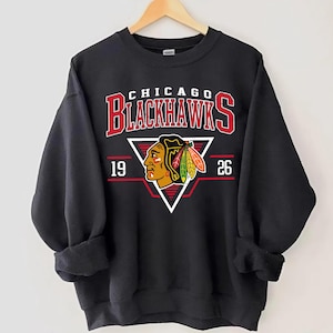 May include: Black sweatshirt with a vintage-style graphic of the Chicago Blackhawks logo. The logo features a red and black Native American headdress with a feather and the text "Chicago Blackhawks" in red and black. The numbers "19" and "26" are on either side of the logo.