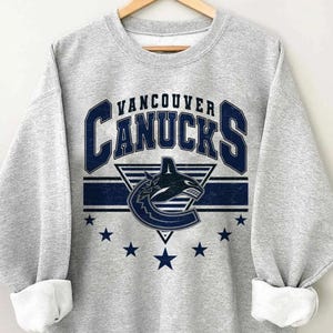 Pu&ograve; includere: Una felpa grigio melange con la scritta "Vancouver Canucks" in blu navy, il logo della squadra e stelle. La felpa &egrave; esposta su una gruccia di legno.