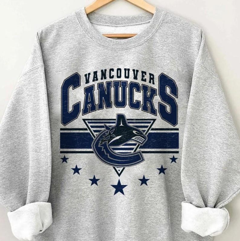 Pu&ograve; includere: Una felpa grigio melange con la scritta "Vancouver Canucks" in blu navy, il logo della squadra e stelle. La felpa &egrave; esposta su una gruccia di legno.