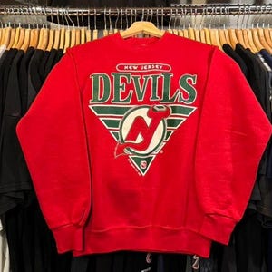 May include: Red sweatshirt with a green, white and red New Jersey Devils logo. The logo features the team's name and a stylised 'N' with a hockey stick.