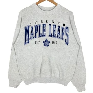 May include: Gray sweatshirt with the words "Toronto Maple Leafs" in blue and white lettering. The logo for the Toronto Maple Leafs is in the center of the sweatshirt. The text "Est. 1917" is below the logo.