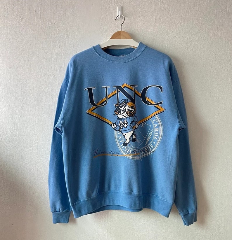K&ouml;nnte beinhalten: Ein hellblaues Sweatshirt mit einem gro&szlig;en Grafikdruck des UNC-Maskottchens, einem Widder, der einen Hut tr&auml;gt und einen Baseballschl&auml;ger h&auml;lt. Die Grafik enth&auml;lt auch den Text "UNC" und "University of North Carolina at Chapel Hill".