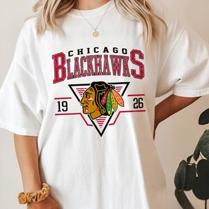 May include: White t-shirt with a red, white, and black Chicago Blackhawks logo. The logo features a Native American headdress with feathers and the text "Chicago Blackhawks" and "19 26".