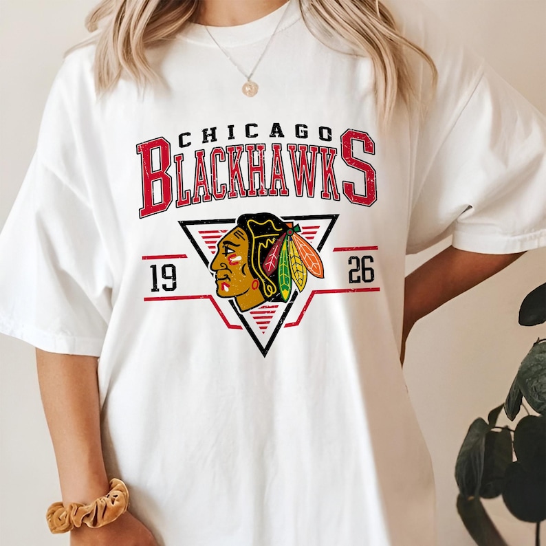 May include: White t-shirt with a red, white, and black Chicago Blackhawks logo. The logo features a Native American headdress with feathers and the text "Chicago Blackhawks" and "19 26".