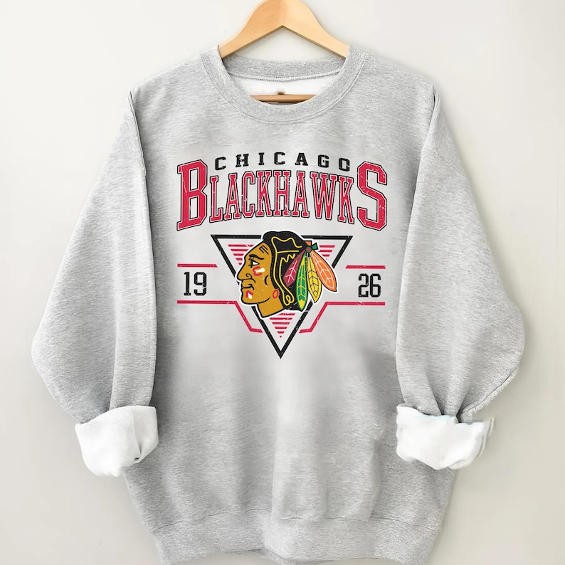 May include: Gray sweatshirt with a vintage-style Chicago Blackhawks logo. The logo features a Native American head with feathers and the text "Chicago Blackhawks" with the years "19" and "26" on either side.