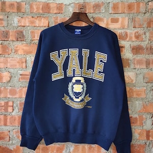May include: A navy blue sweatshirt with a gold glitter Yale University logo. The logo features the school's crest and the words "Yale" and "New Haven".