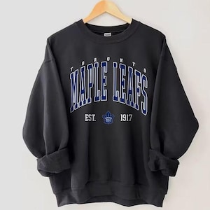 May include: Black crewneck sweatshirt with the text "TORONTO MAPLE LEAFS" in white and blue lettering. Below the text is "EST. 1917" and a maple leaf logo. The sweatshirt is hanging on a wooden hanger.