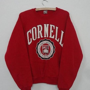 May include: Red sweatshirt with a white Cornell University logo. The logo features a crest with a book, a building, and the text "Cornell University" and "Founded by Ezra Cornell".
