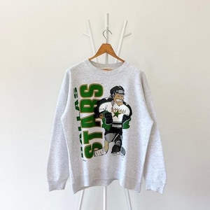 May include: A gray sweatshirt with a green and white Dallas Stars logo. The logo features a cartoon hockey player wearing a green jersey with a white star and the word "Stars" on it.