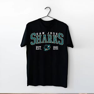 May include: Black t-shirt with a white graphic of the San Jose Sharks logo. The logo includes the team name "San Jose Sharks" and the text "Est. 1991".