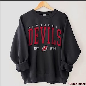 May include: Black sweatshirt with the text "NEW JERSEY DEVILS" in red block letters. Below the text is "EST. 1974" and a red and white logo. The sweatshirt is hanging on a wooden hanger.