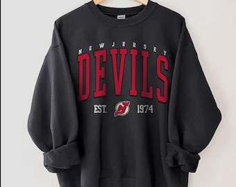 May include: Black sweatshirt with the text "NEW JERSEY DEVILS" in red block letters. Below the text is "EST. 1974" and a red and white logo. The sweatshirt is hanging on a wooden hanger.