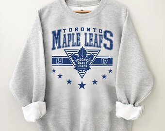 May include: A light gray sweatshirt with the text "TORONTO MAPLE LEAFS" in blue. The design includes a maple leaf emblem and the numbers "19" and "17". Blue star accents are below the text. The sleeves are cuffed.
