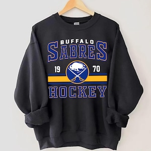 May include: Black sweatshirt with a vintage-style Buffalo Sabres hockey logo. The logo features a blue and gold buffalo head with crossed hockey sticks and the text "Buffalo Sabres" in a blue and gold font. The text "19 70" is below the logo, and the text "Hockey" is below that.