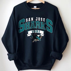 May include: Navy blue sweatshirt with the San Jose Sharks logo. The design features "SAN JOSE SHARKS 1991" in white and teal, plus a shark logo. The sweatshirt has a crew neck and long sleeves, perfect for cooler weather.
