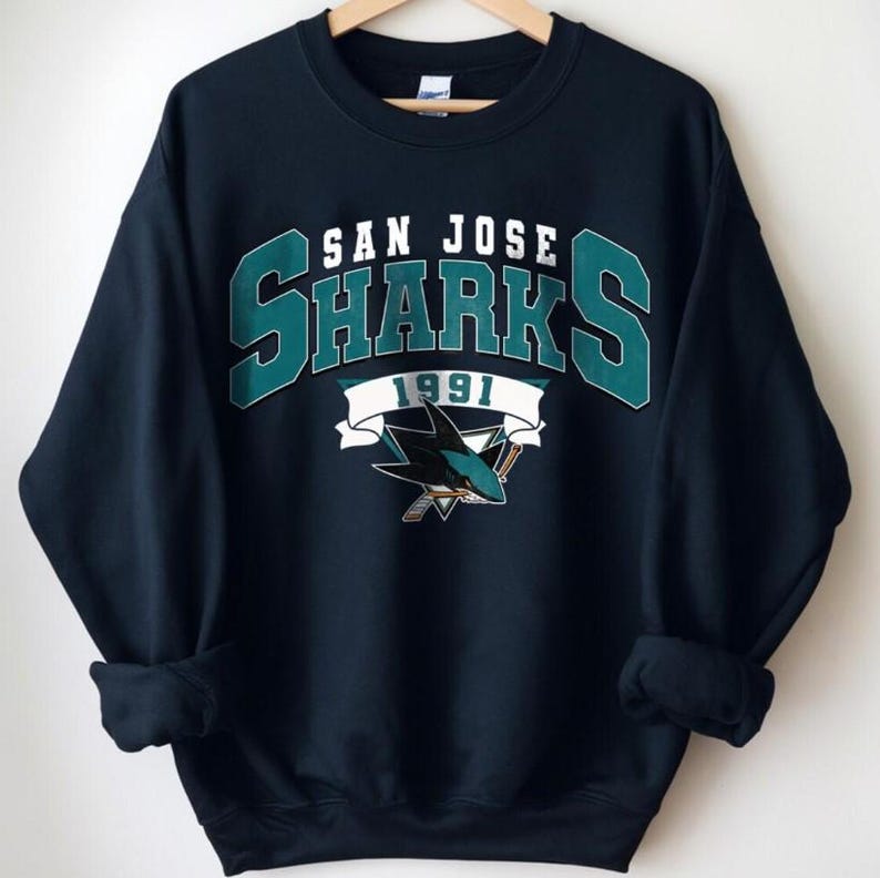May include: Navy blue sweatshirt featuring the San Jose Sharks logo. The design includes the words "SAN JOSE SHARKS 1991" in white and teal lettering, along with a shark logo. The sweatshirt has a crew neck and long sleeves.