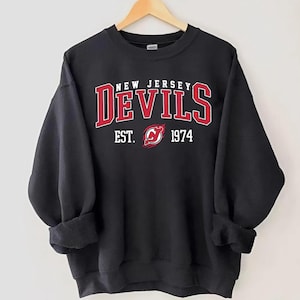 May include: Black sweatshirt with the text "NEW JERSEY DEVILS" in red and white lettering. The sweatshirt also features the team logo and the year "1974". The sleeves are rolled up.