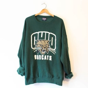 May include: Green sweatshirt with a white and gold Ohio Bobcats logo. The logo features a cartoon wildcat with the word "Ohio" above it and "Bobcats" below it.